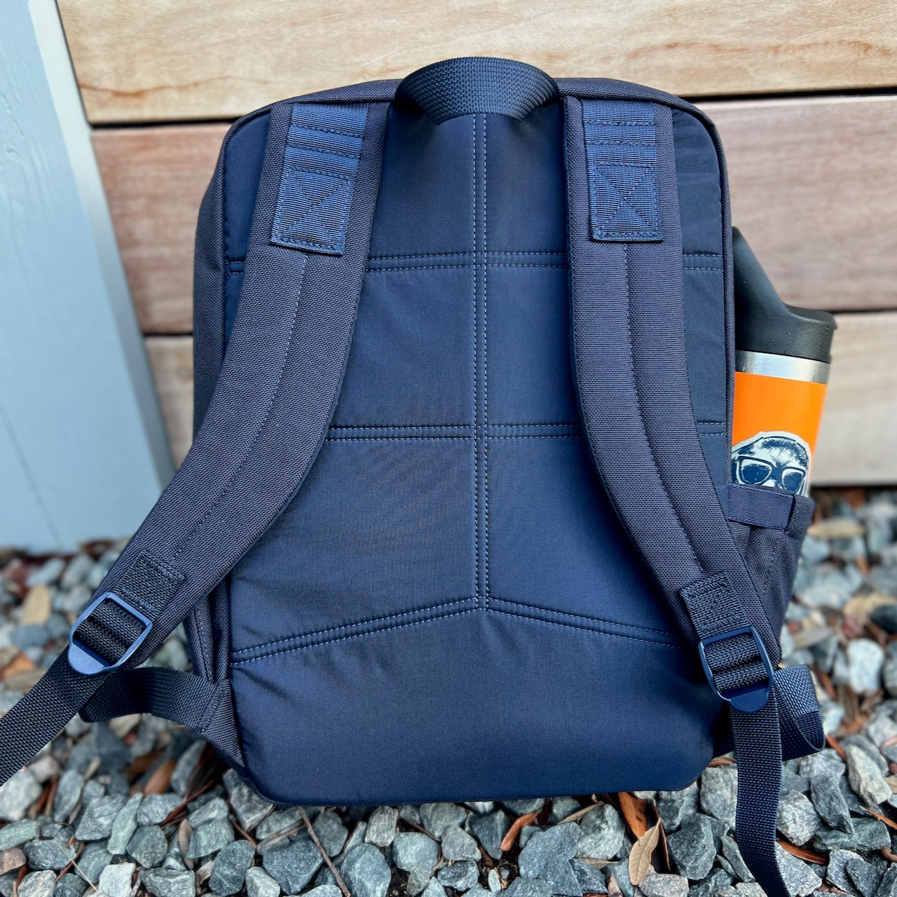 The KR1 2.0 - Kid Ruck - Add Patches, Make it Yours backpack in navy blue rests on gravel, featuring a colorful water bottle in its side pocket—a durable kids backpack designed for every adventure.