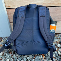 The KR1 2.0 - Kid Ruck - Add Patches, Make it Yours backpack in navy blue rests on gravel, featuring a colorful water bottle in its side pocket—a durable kids backpack designed for every adventure.