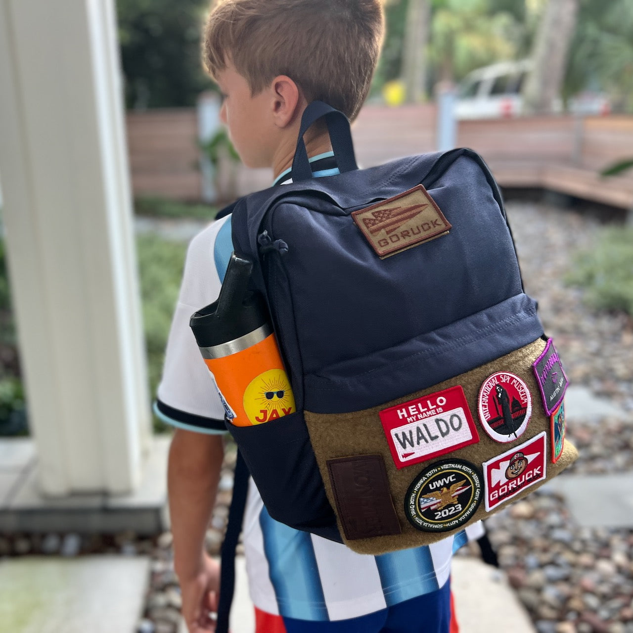 A boy stands outside on a stone path, wearing the KR1 2.0 - Kid Ruck backpack adorned with vibrant, customizable patches.