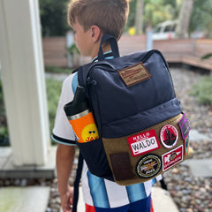 A boy stands outside on a stone path, wearing the KR1 2.0 - Kid Ruck backpack adorned with vibrant, customizable patches.