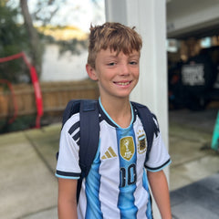 A smiling boy stands outside near a garage, wearing an Argentina soccer jersey and carrying the KR1 2.0 - Kid Ruck backpack, ready to add patches and make it his own.