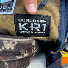A “KR1 2.0 - Kid Ruck” patch on tan fabric, with a finger pointing above it—showcasing the reliable quality you expect from an ultra-tough everyday book bag that kids can customize with their own patches.