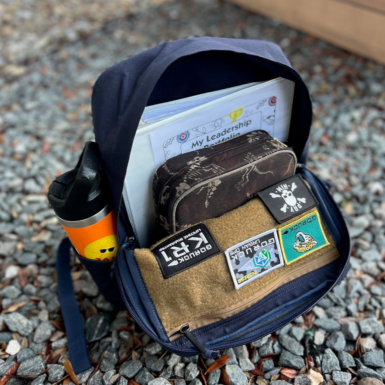 The KR1 2.0 - Kid Ruck - Add Patches, Make it Yours is open on gravel, displaying folders, a water bottle, a brown pouch, and sewn-on patches inside.