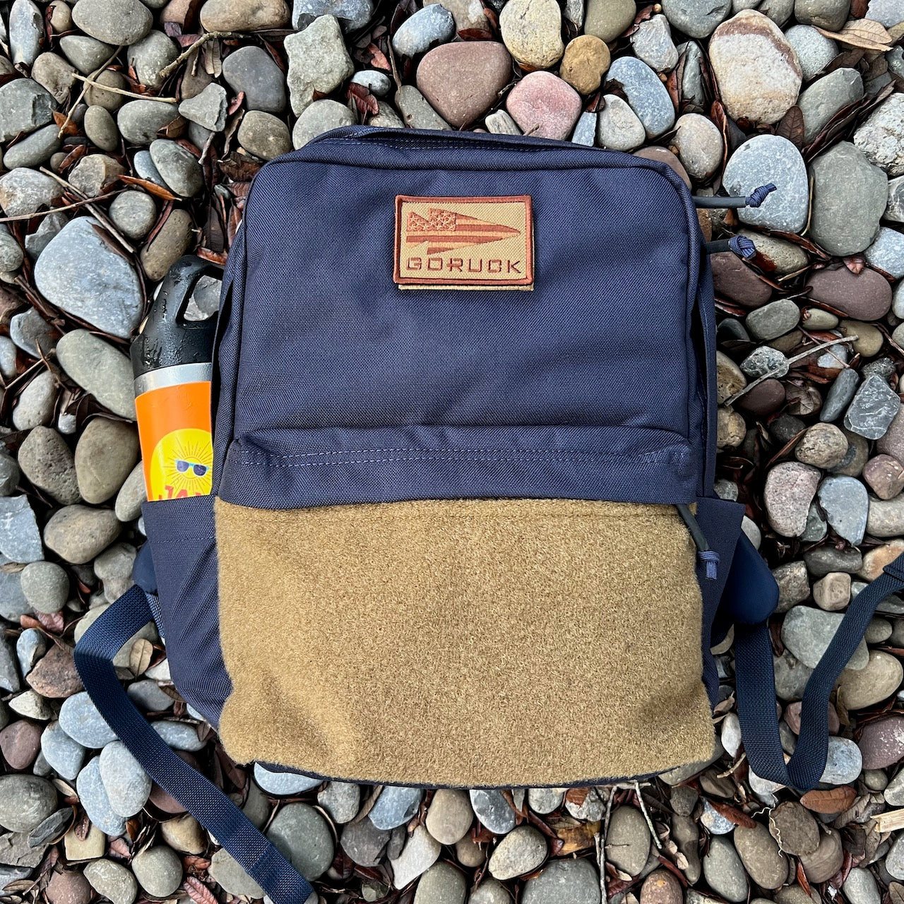 A KR1 2.0 - Kid Ruck backpack in blue and brown, designed for durability and customizable with patches, along with a water bottle, rests on gray and brown rocks.