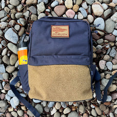 A KR1 2.0 - Kid Ruck backpack in blue and brown, designed for durability and customizable with patches, along with a water bottle, rests on gray and brown rocks.