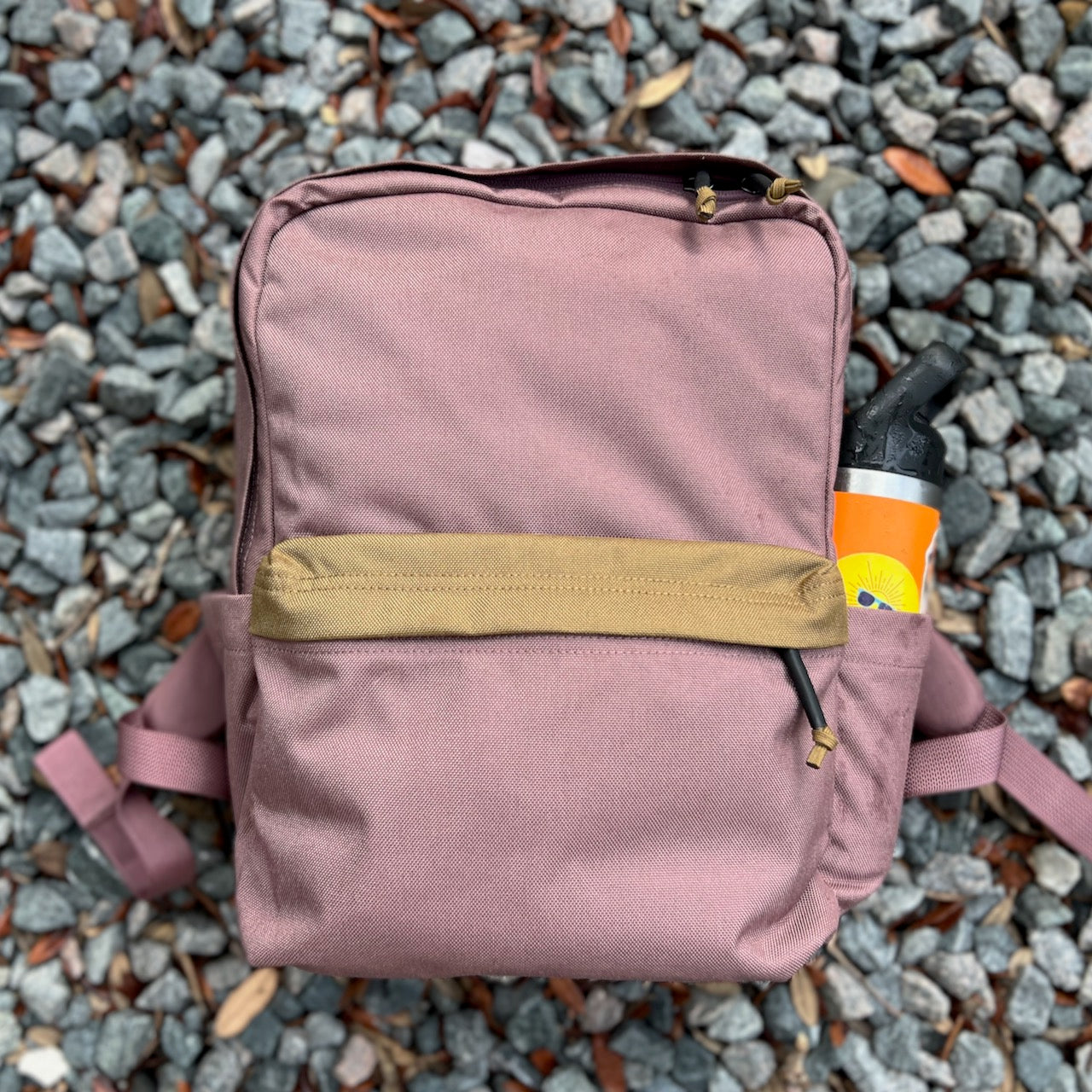 A mauve KR1 2.0 - Kid Ruck backpack with a tan pocket rests on gravel, carrying an orange water bottle in its side pocket—this durable kids backpack is built to handle every adventure and lets you add patches to make it yours.