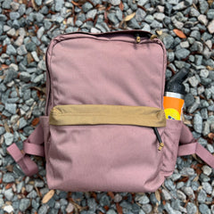 A mauve KR1 2.0 - Kid Ruck backpack with a tan pocket rests on gravel, carrying an orange water bottle in its side pocket—this durable kids backpack is built to handle every adventure and lets you add patches to make it yours.