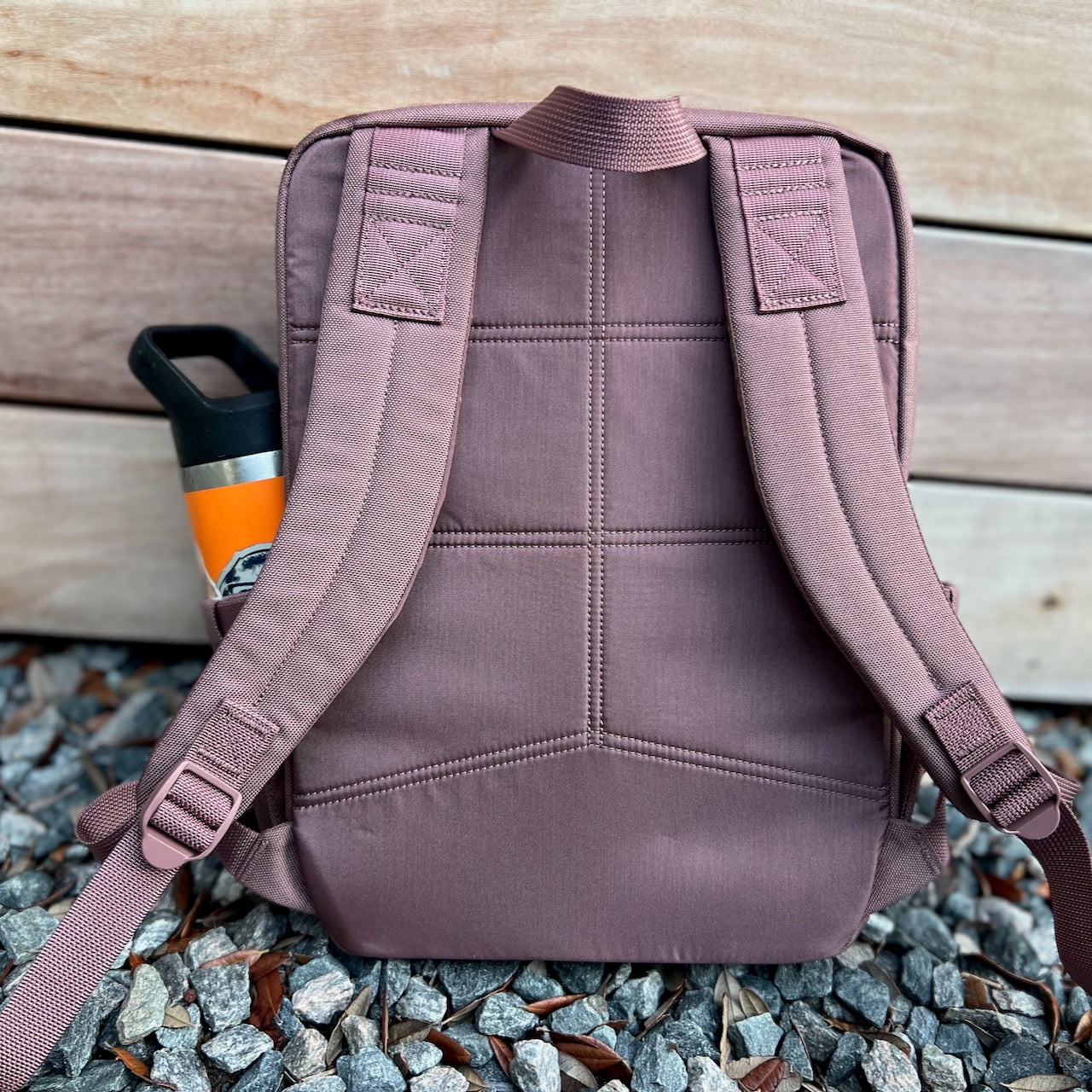 The KR1 2.0 - Kid Ruck backpack, featuring padded straps and designed for kids, sits on gravel with an orange water bottle tucked into its side pocket.