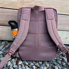 The KR1 2.0 - Kid Ruck backpack, featuring padded straps and designed for kids, sits on gravel with an orange water bottle tucked into its side pocket.