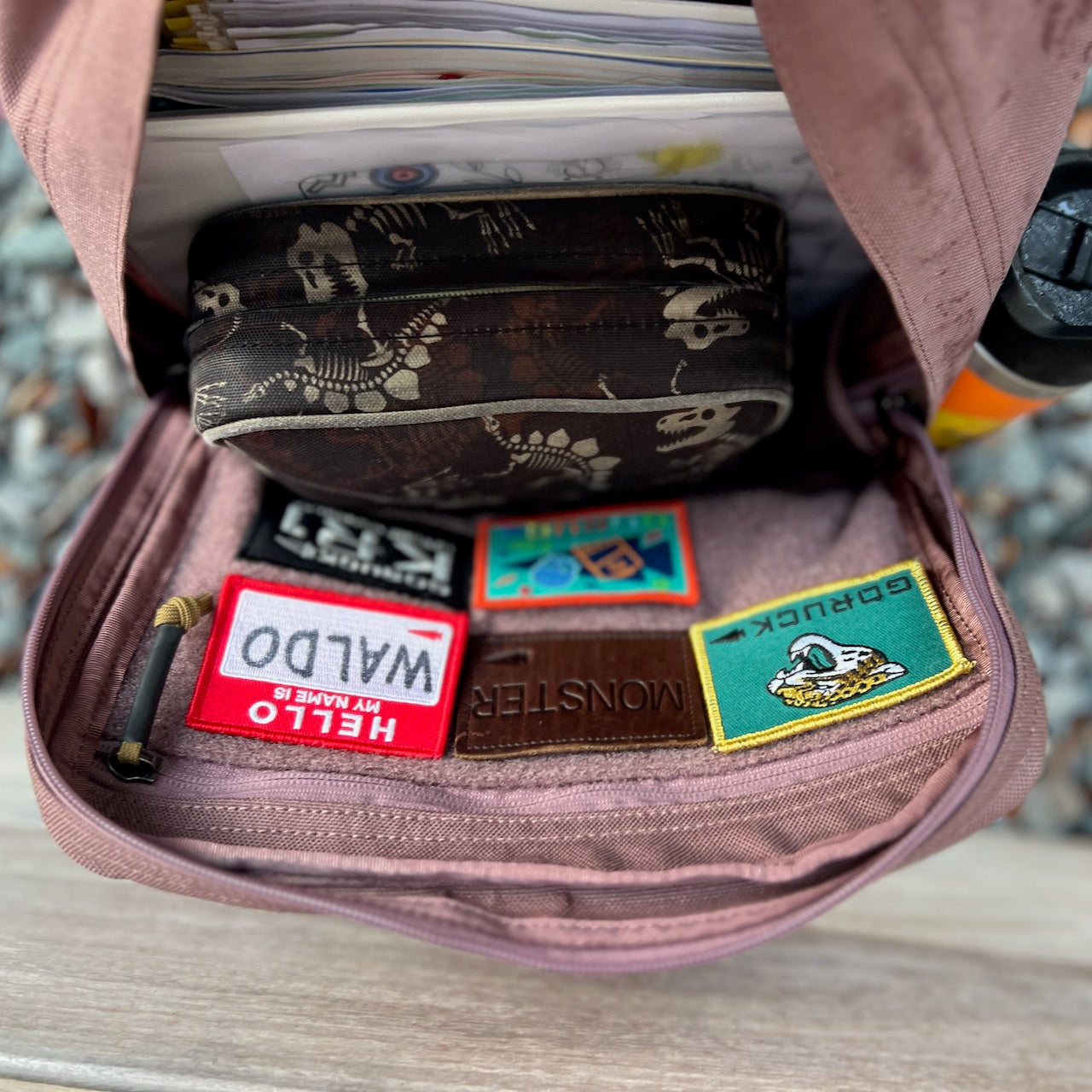 Open the pink KR1 2.0 - Kid Ruck—with custom patches—along with a skull-patterned pouch, papers, and a water bottle on a wooden bench.