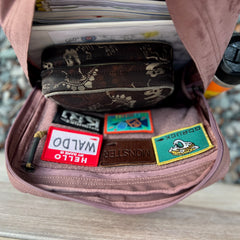 Open the pink KR1 2.0 - Kid Ruck—with custom patches—along with a skull-patterned pouch, papers, and a water bottle on a wooden bench.