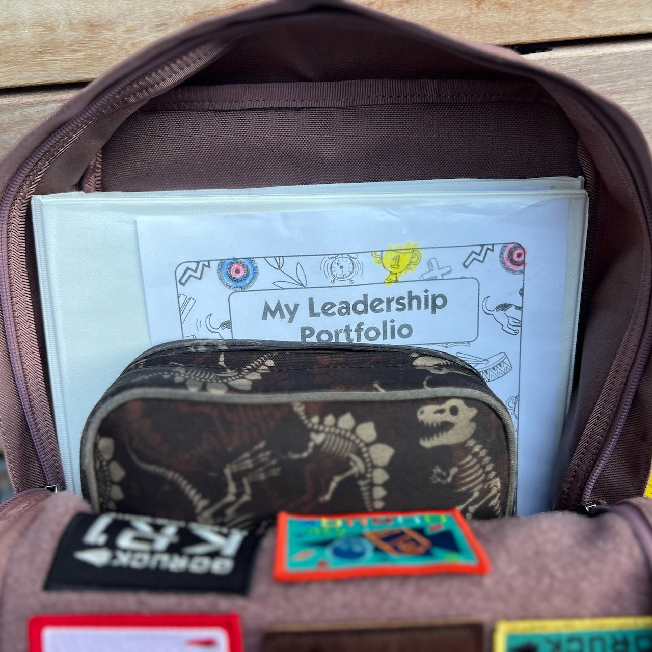 An open, durable KR1 2.0 - Kid Ruck backpack reveals a dinosaur case and a folder labeled “My Leadership Portfolio” inside.
