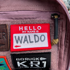 A red “Hello my name is Waldo” patch rests on a fabric surface alongside other patches and a zipper, bringing playful personality to the KR1 2.0 - Kid Ruck - Add Patches, Make it Yours backpack—ideal for customizing your indestructible book bag.