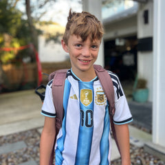 A smiling boy stands outdoors wearing an Argentina soccer jersey and carrying the KR1 2.0 - Kid Ruck, a pink, durable kids backpack you can customize with patches to make it yours.