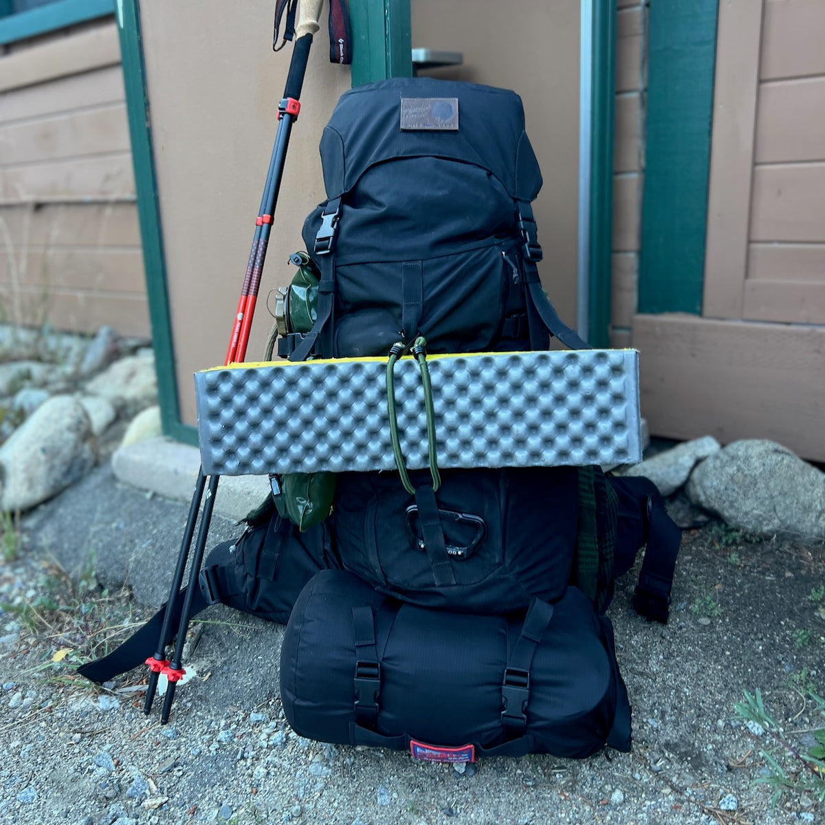 A black climbing-certified hiking backpack with a folded foam sleeping pad attached leans against a wall next to a pair of trekking poles and an aluminum carabiner.