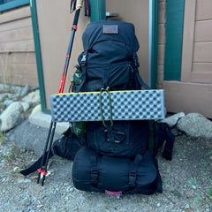A black climbing-certified hiking backpack with a folded foam sleeping pad attached leans against a wall next to a pair of trekking poles and an aluminum carabiner.