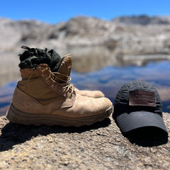 The MACV-2 Mid Top boots in Briar + Coyote and a black “MONSTER” cap sit atop a lakeside rock with mountains in the background, evoking the rugged design of classic Special Forces footwear such as the legendary Vietnam jungle boot.