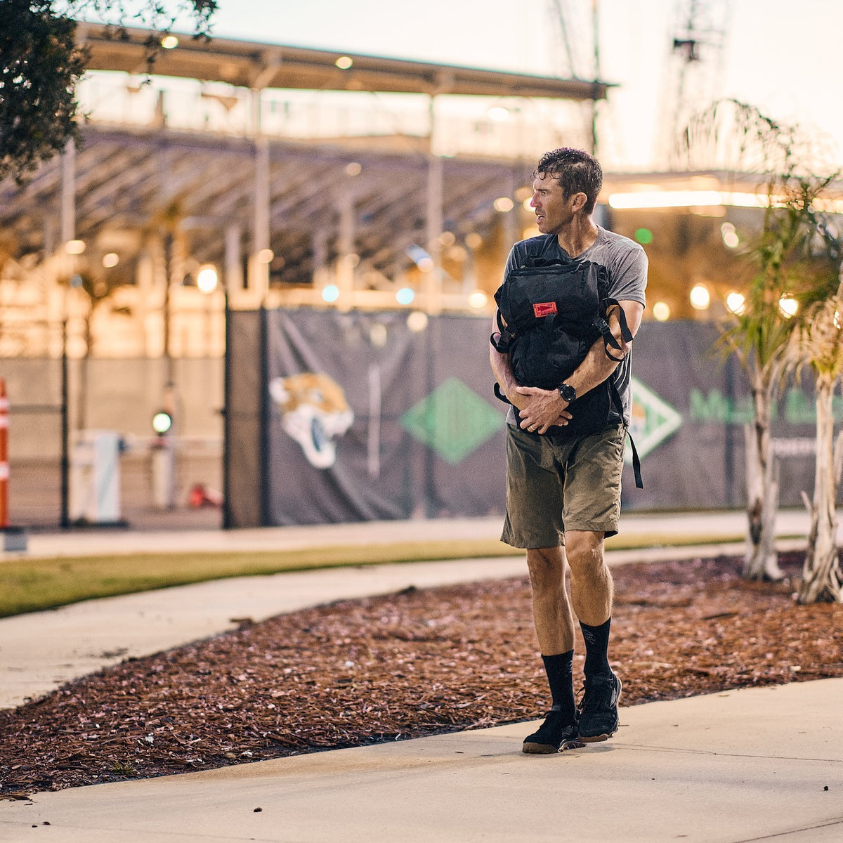 A man wearing a backpack and tough shorts made of 2-way stretch fabric walks along a sidewalk near a stadium.