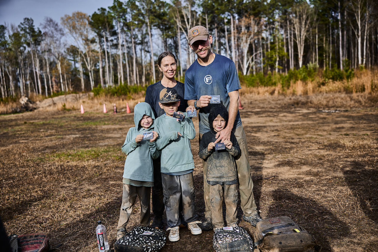 A muddy family of five smiles and holds cards after completing the BRAGG 2027 event in a wooded area, celebrating a challenge inspired by Special Forces training.