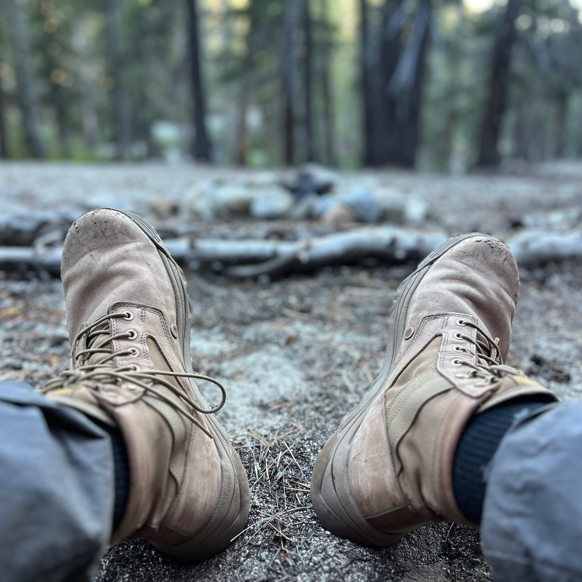 A person wearing MACV-2 - Mid Top - Briar + Coyote boots sits on the forest ground, with trees and rocks softly blurred in the background.