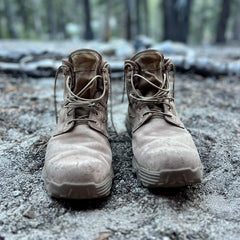 A pair of MACV-2 - Mid Top - Briar + Coyote boots sit on dirt in a forest, with blurred trees in the background.