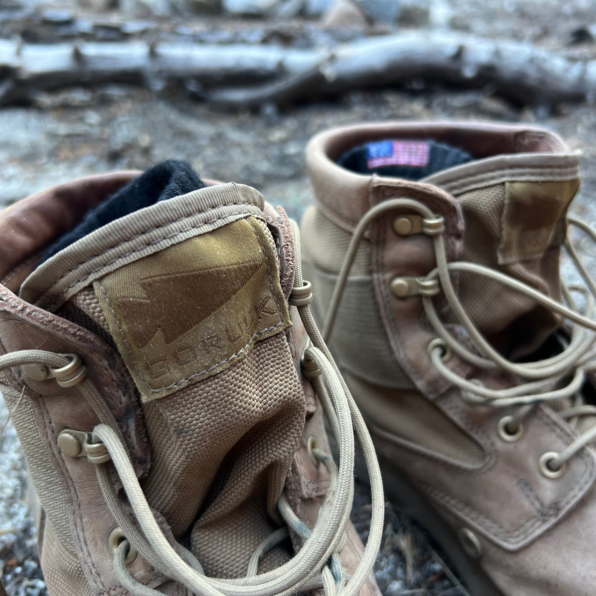 A close-up of tan MACV-2 Mid Top boots in Briar + Coyote with untied laces on a rocky outdoor surface.