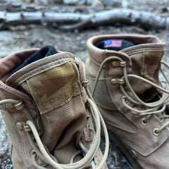 A close-up of tan MACV-2 Mid Top boots in Briar + Coyote with untied laces on a rocky outdoor surface.