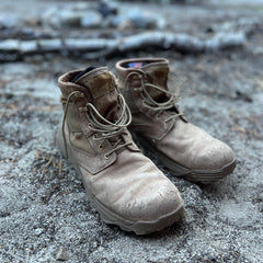 A pair of MACV-2 - Mid Top - Briar + Coyote boots rests on sandy ground outdoors, with branches in the background.