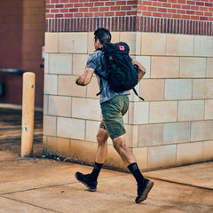 A person in a grey t-shirt, tough green shorts, and a black backpack runs past a brick building.