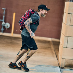 A man in athletic wear and tattoos runs outdoors near a brick wall with the Mackall 45 Fog—a red patterned backpack featuring a Special Forces rucking chassis.
