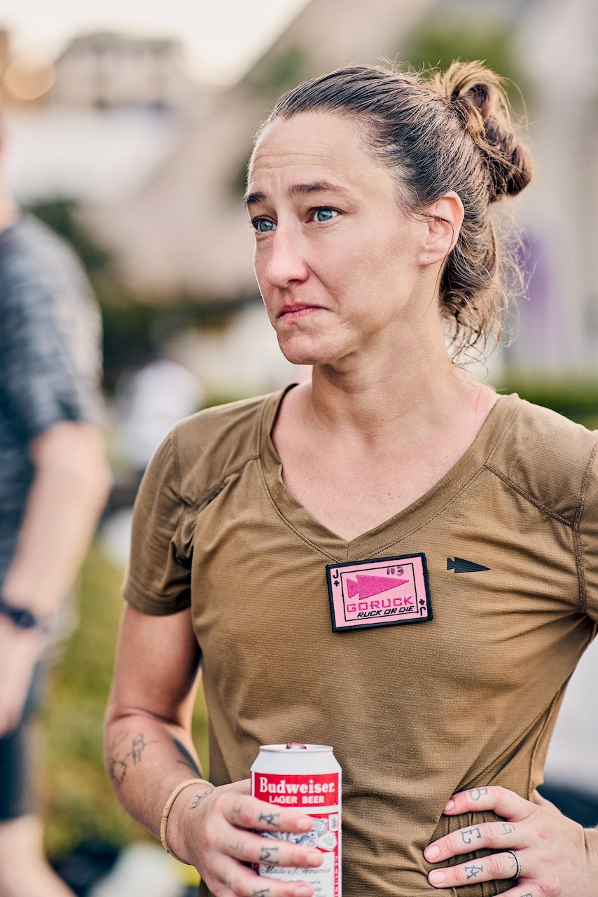 A woman wearing a tan t-shirt with a pink “RUCK OR DIE” patch holds a can of Budweiser at an Austin rucking event.