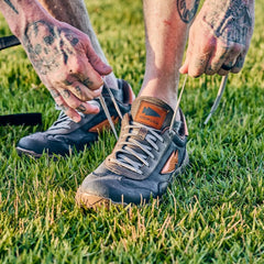 Tattooed hands tying the laces on the Mackall 45 Fog sneaker with a heritage suede upper, while standing on green grass.