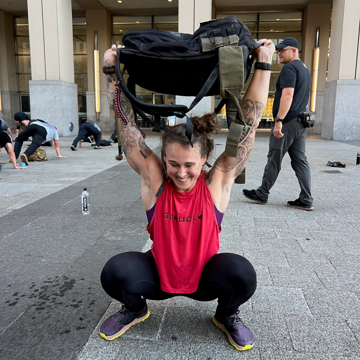 A woman squats outdoors, lifting a heavy backpack overhead in her Women’s Performance Tank - ToughMesh, with others exercising in the background. Made in USA.