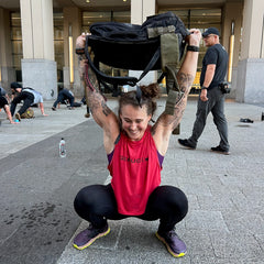 A woman squats outdoors, lifting a heavy backpack overhead in her Women’s Performance Tank - ToughMesh, with others exercising in the background. Made in USA.