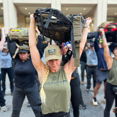 A group of people outdoors lift weighted backpacks overhead, with a woman sporting a blonde braid in front—all wearing athletic gear, including the Women’s Performance Tank - ToughMesh, designed to keep them cool and focused.