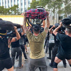 A group of people outdoors, facing away from the camera, lifts backpacks and helmets overhead—several are wearing hi-vis GORUCK Reflective Tee - Tri-Blend shirts reminiscent of Special Forces teams.