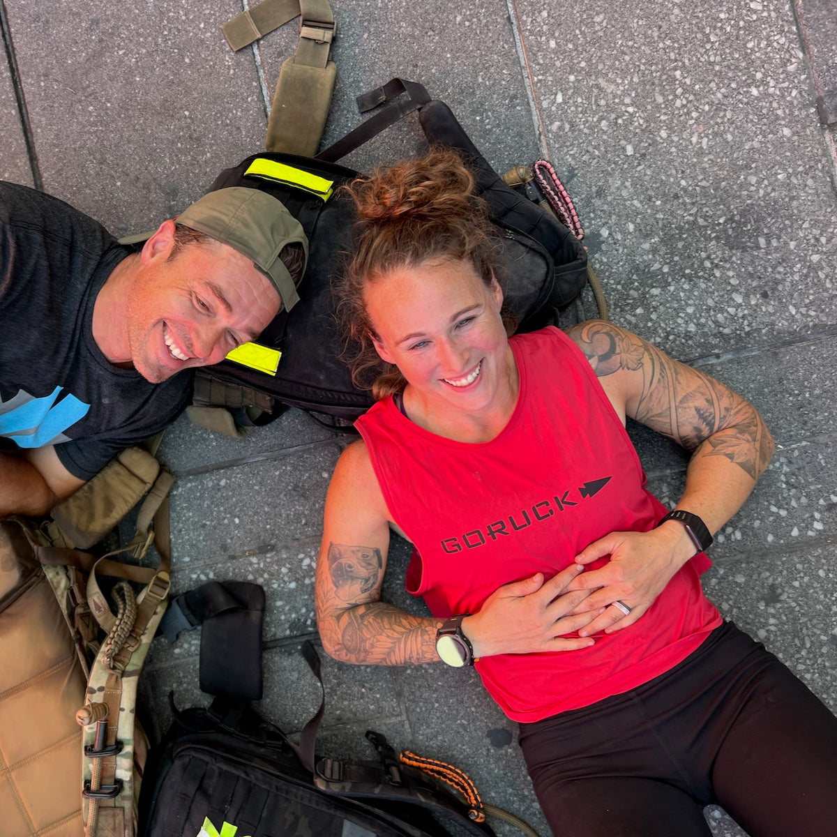 Two people smiling, lying on the ground with backpacks—one wearing the Women’s Performance Tank made from featherweight ToughMesh fabric in red, crafted in the USA by GORUCK, and the other sporting a cap.