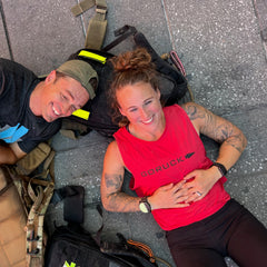Two people smiling, lying on the ground with backpacks—one wearing the Women’s Performance Tank made from featherweight ToughMesh fabric in red, crafted in the USA by GORUCK, and the other sporting a cap.