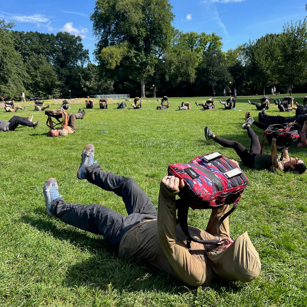 People exercising on grass, lifting their backpacks—each featuring rugged durability—above their chests, with trees and blue sky in the background. The group uses the Mackall 45 - Americana rucking pack to enhance strength and endurance during their outdoor workout.