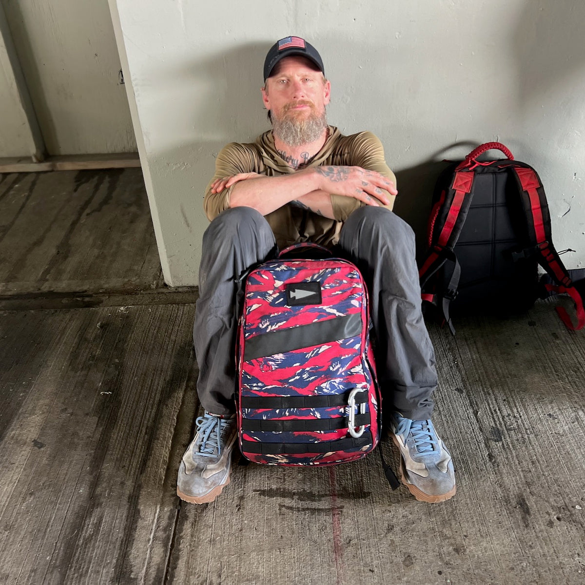 A man sits against a wall, arms resting on a ruggedly durable red-and-black backpack, while the Mackall 45 – Americana, designed for performance worthy of a Special Forces rucking chassis, rests beside him.