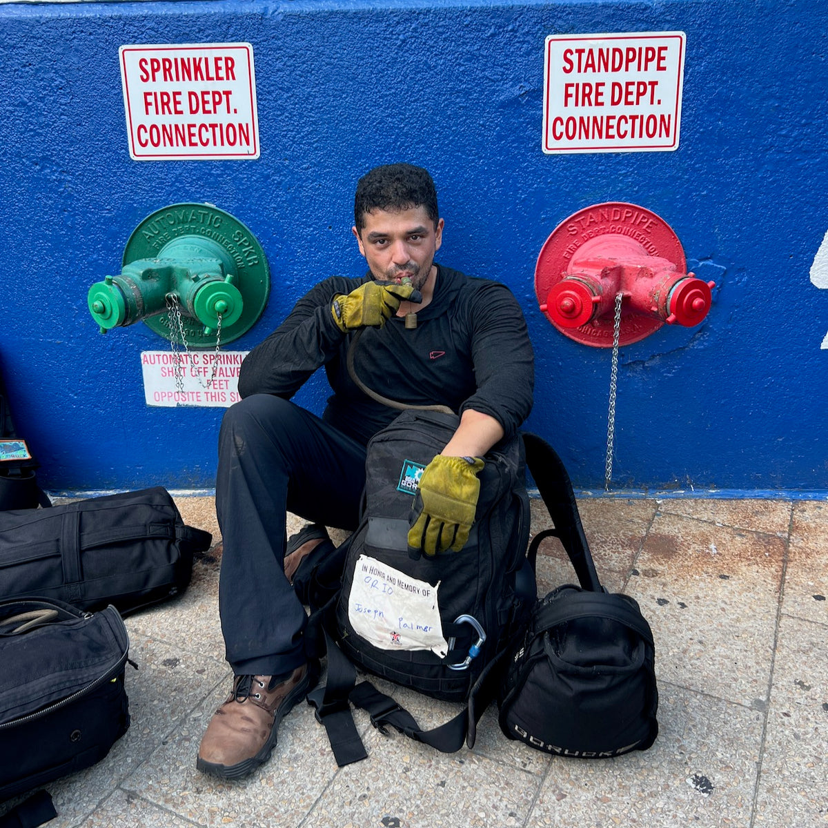 A man wearing MACV-2 - Mid Top - Chestnut + Black boots sits by green and red fire hydrant connections, holding a hose, with bags on the ground beside him.