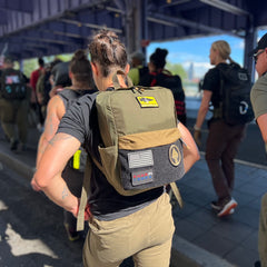 Wearing the KR1 2.0 - Kid Ruck - Add Patches, Make it Yours—a durable kids backpack designed for personalization and backed by a lifetime guarantee—a child walks with a group under a shaded walkway on a sunny day.