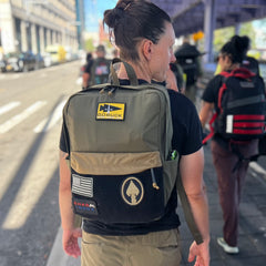 A person carrying the KR1 2.0 - Kid Ruck backpack, customizable with patches, walks along a city sidewalk with others in the background.