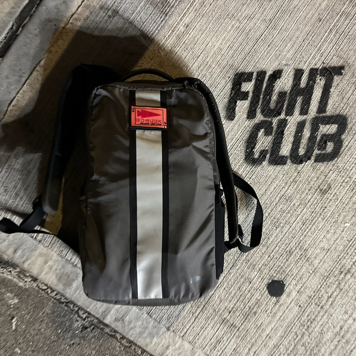 A Basic Rucker backpack in gray, featuring a red patch, rests on concrete beside the bold, black stenciled words “FIGHT CLUB,” embodying the rugged essence of rucking with this popular GORUCK product.