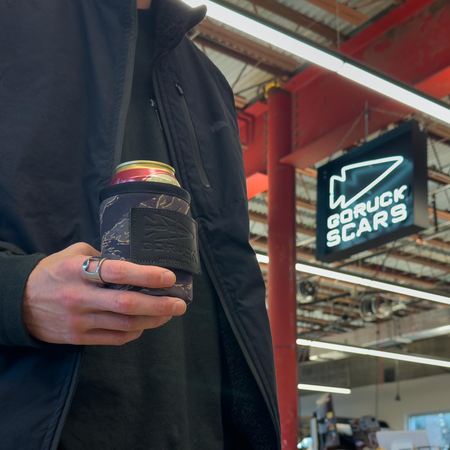 A person holds a canned drink in a Ballistic Beer Jacket - GORUCK, standing indoors near a GORUCK SCARS sign.