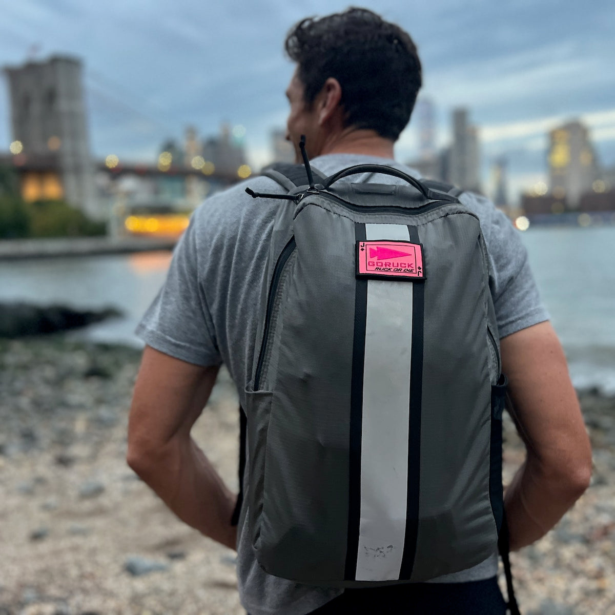 A man with short hair, wearing a gray shirt and carrying the Basic Rucker backpack, stands near a rocky waterfront with views of the city skyline.