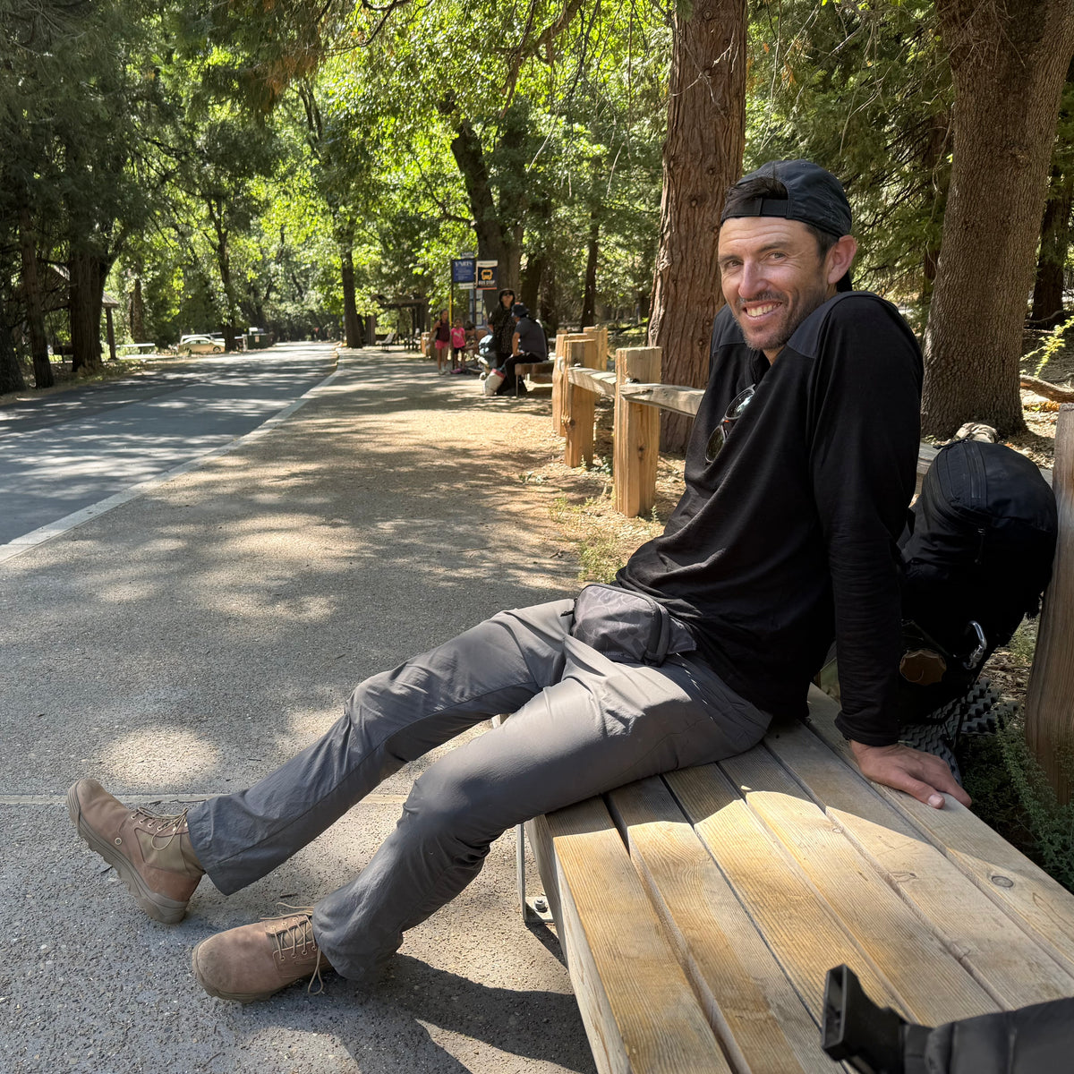 A man in hiking gear wearing MACV-2 Mid Top boots in Briar + Coyote relaxes on a wooden bench by a forested trail on a sunny day.