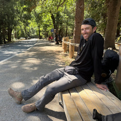 A man in hiking gear wearing MACV-2 Mid Top boots in Briar + Coyote relaxes on a wooden bench by a forested trail on a sunny day.