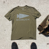 Olive green GORUCK Reflective Tee - Tri-Blend on concrete beside a bag, with black shoes in frame—highlighting Special Forces-inspired durability and style.