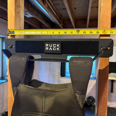 A wall-mounted tactical gear organization rack sits between two wooden studs, measured by a yellow tape measure showing a width of approximately 16 inches.